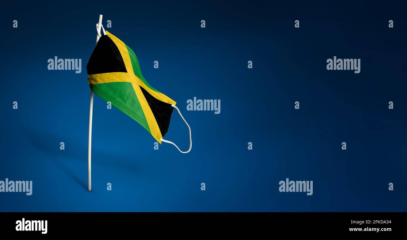 Maschera Giamaica su sfondo blu scuro. Bandiera ondulata della Giamaica dipinta su maschera medica sul palo. Concetto del banner della lotta contro l'epidemia Foto Stock