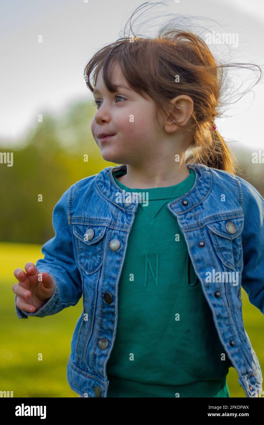 Ritratto di una bambina carina, dai capelli castani, dagli occhi blu, con giacca blu e ponticello verde che corre in un parco in una giornata di sole Foto Stock