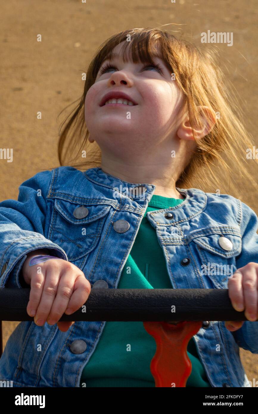 Una ragazza carina, dai capelli marroni, dagli occhi blu, che indossa una giacca blu e un ponticello verde seduto in un'altalena in una giornata di sole primavera Foto Stock