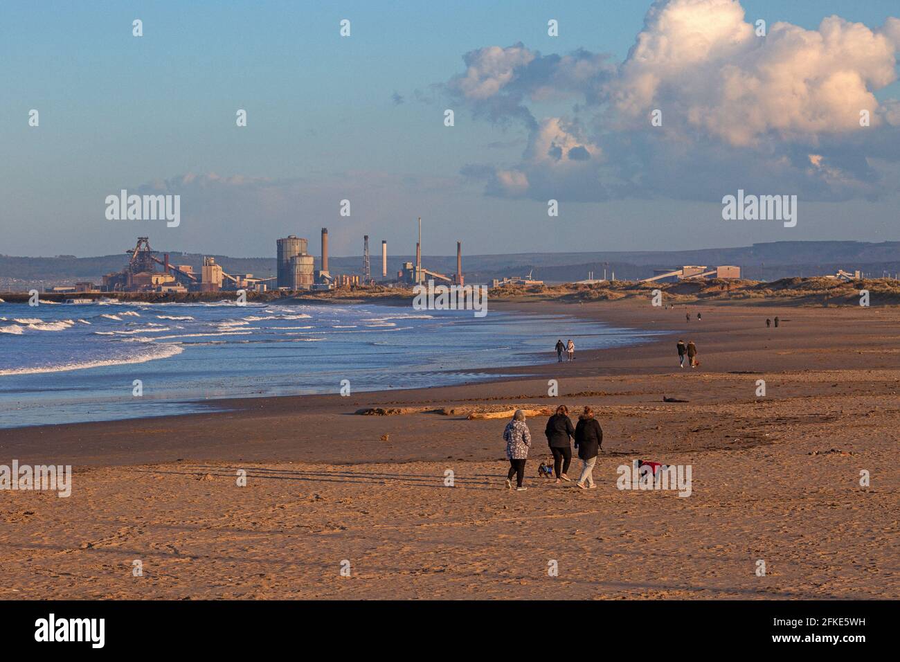 Persone che camminano lungo la spiaggia con le acciaierie Redcar in distanza, Inghilterra, Regno Unito Foto Stock