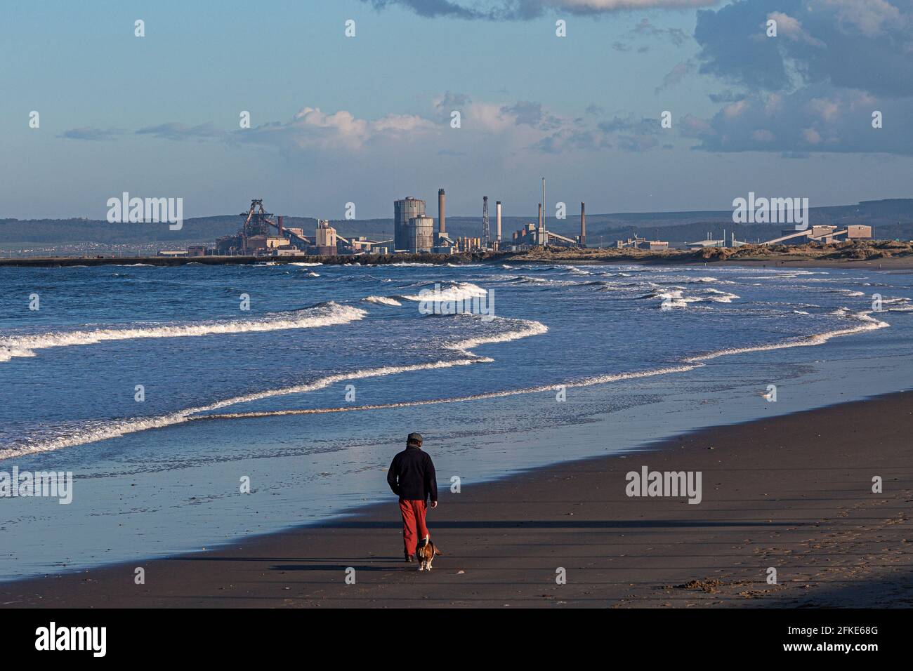 Uomo e il suo vecchio cane camminando lungo la spiaggia con le acciaierie Redcar in distanza, Inghilterra, Regno Unito Foto Stock