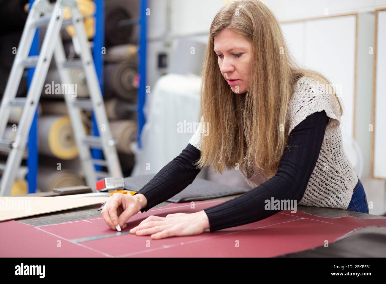Una lavoratrice che prepara e contrassegna un pezzo di pelle in un'officina di tappezzeria. È bionda e caucasica. Foto Stock