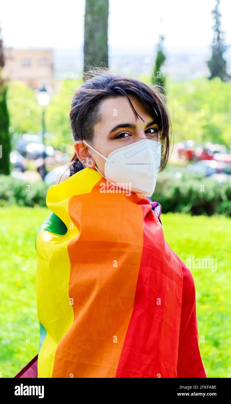 Una transgender / non ragazza con la bandiera gay sulle spalle e una maschera chirurgica. Persone autentiche non binarie. Gay Pride Day durante la pandemia di Covid Foto Stock