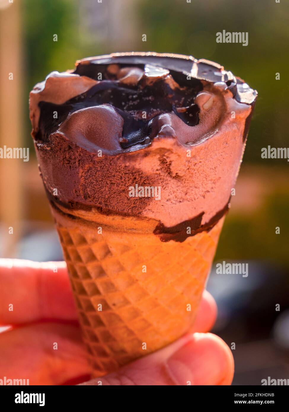 Cono gelato al cioccolato tenuto contro un verde e parcheggiato le auto si sono offuscate sullo sfondo Foto Stock