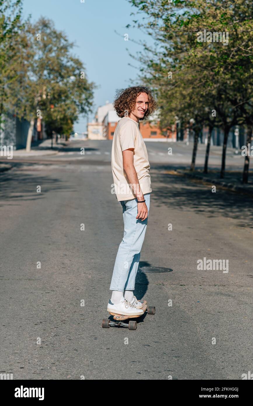 Uomo caucasico dai capelli rossi che usa uno skateboard. Indossa abiti casual. Pattina lungo la strada di una città circondata da alberi. Giornata estiva soleggiato con har Foto Stock