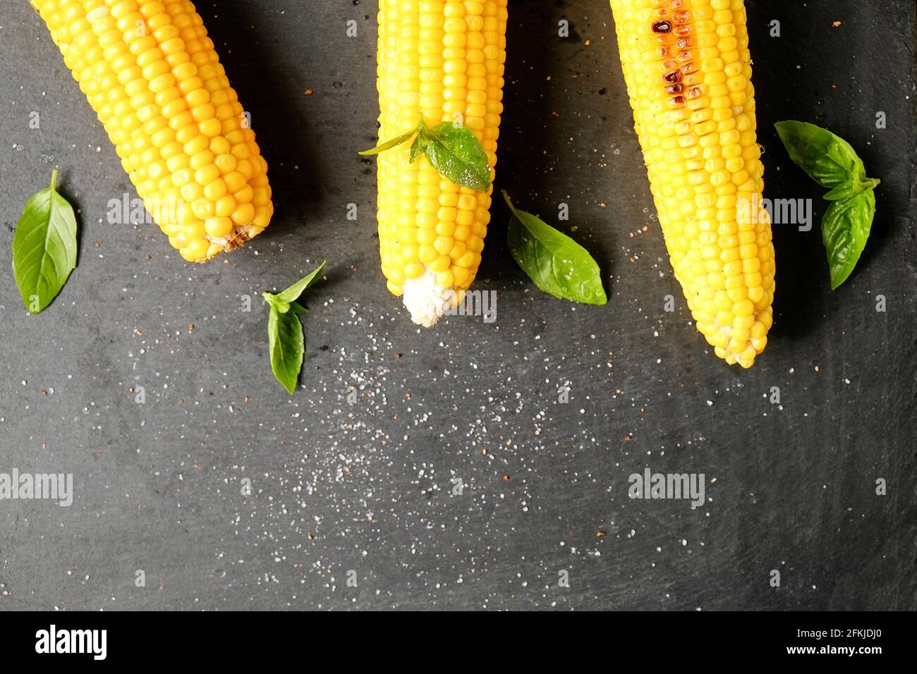 Tre pannocchie di mais grigliate sul fuoco al barbecue posate con foglie di basilico verde e decorazione di sale su tavola di pietra scura. Concetto di abitudini alimentari pulite. Backgro Foto Stock
