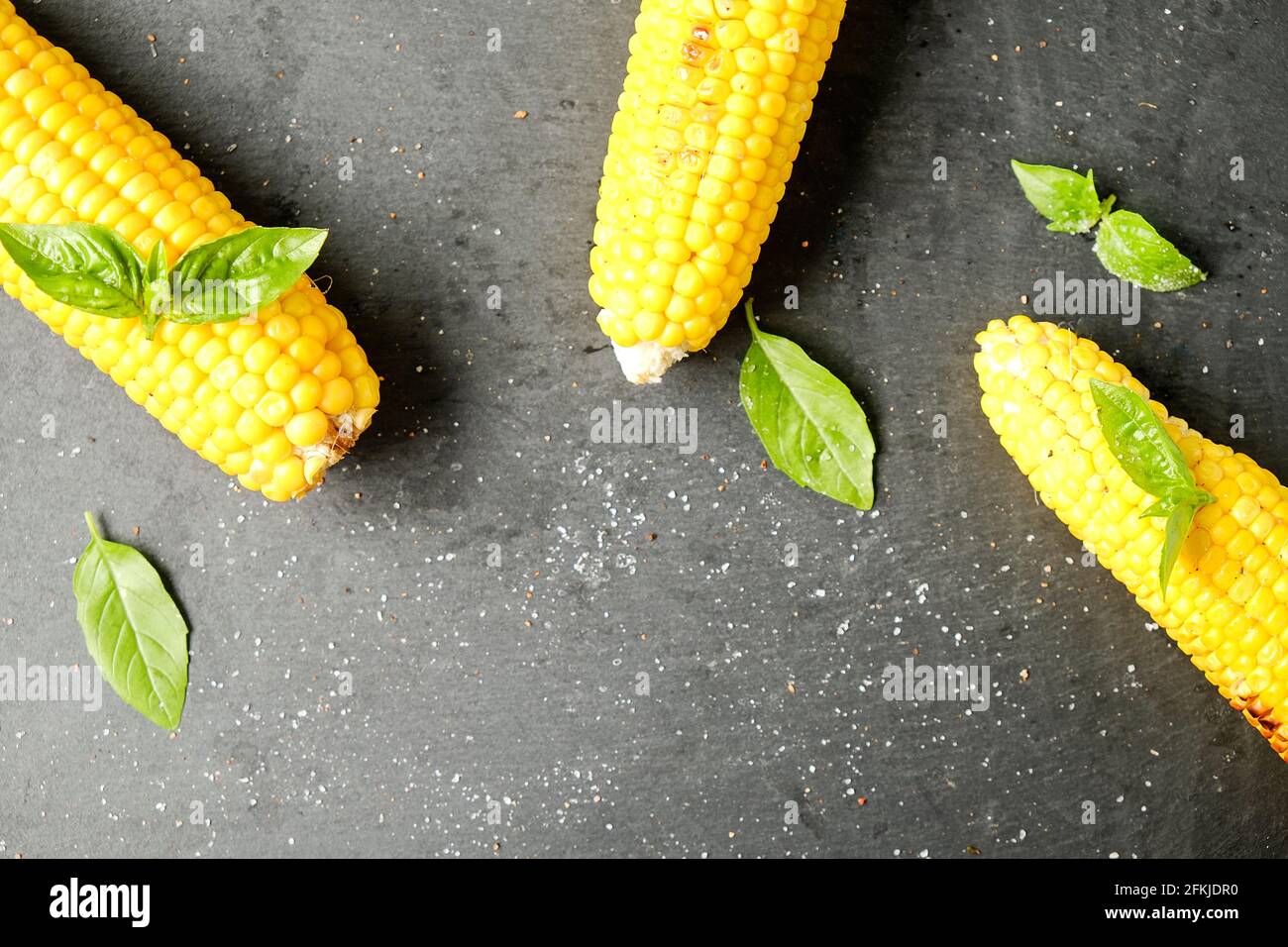 Tre pannocchie di mais grigliate sul fuoco al barbecue posate con foglie di basilico verde e decorazione di sale su tavola di pietra scura. Concetto di abitudini alimentari pulite. Backgro Foto Stock