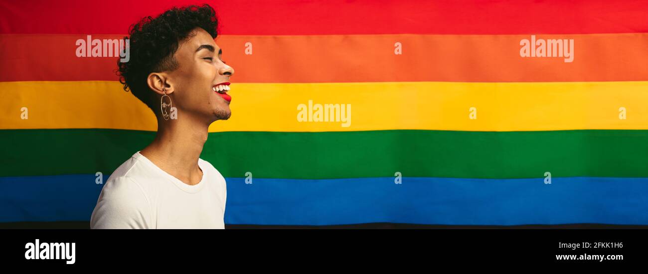 Vista del profilo di gay uomo in piedi contro bandiera di orgoglio e sorridente. Banner di un uomo transgender che indossa orecchini e trucco agasint una bandiera arcobaleno. Foto Stock