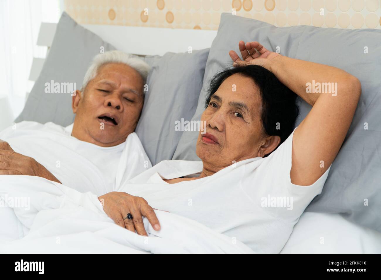 La donna asiatica più anziana che si stese gli occhi a letto accanto al marito che russava e fa rumore, i problemi dei coniugi. Foto Stock
