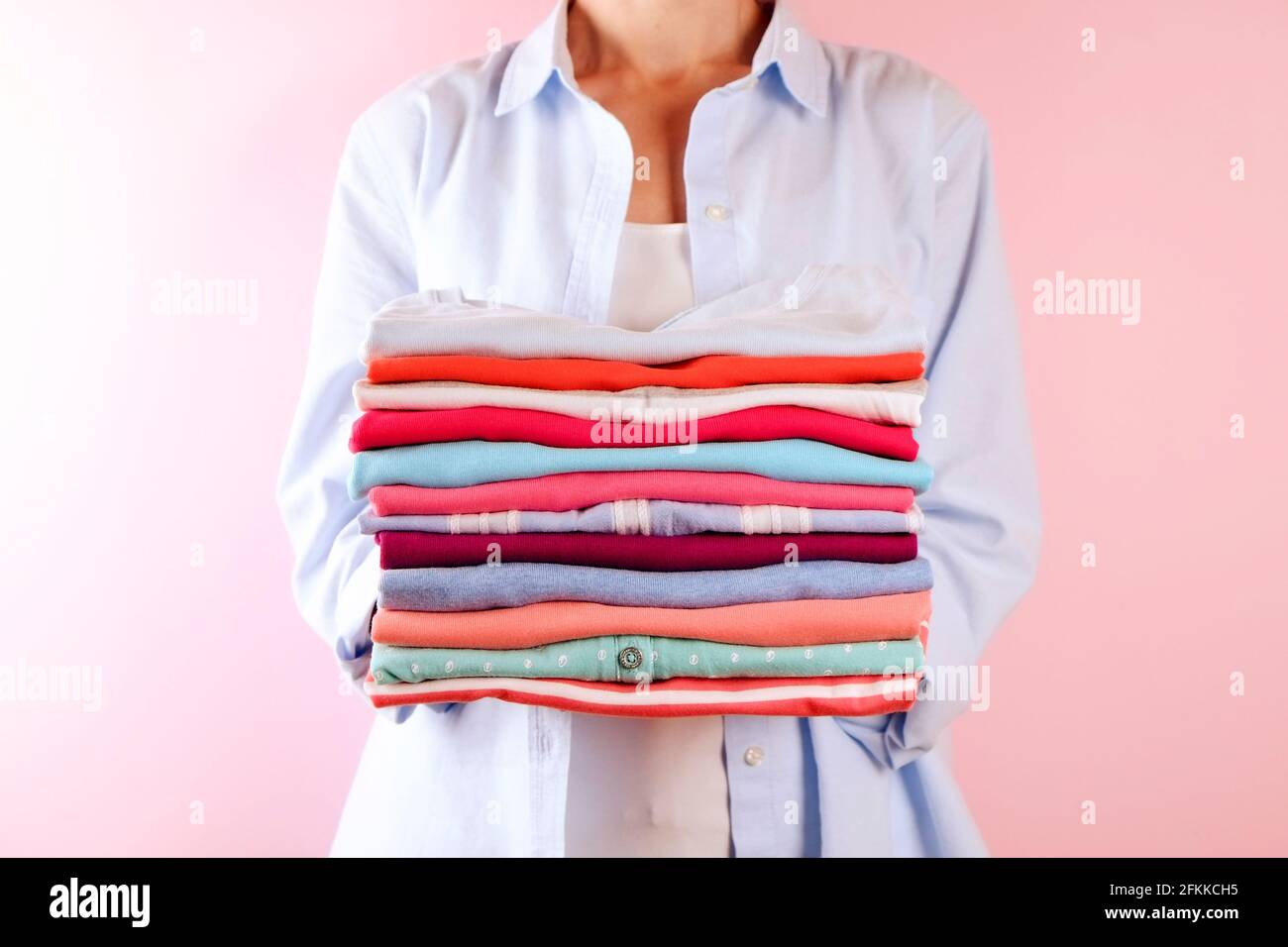 Primo piano di una giovane donna che indossa una camicia di cotone blu con una pila di camicie multicolore perfettamente piegate. Femmina con pile di indumenti di colore diverso in h Foto Stock