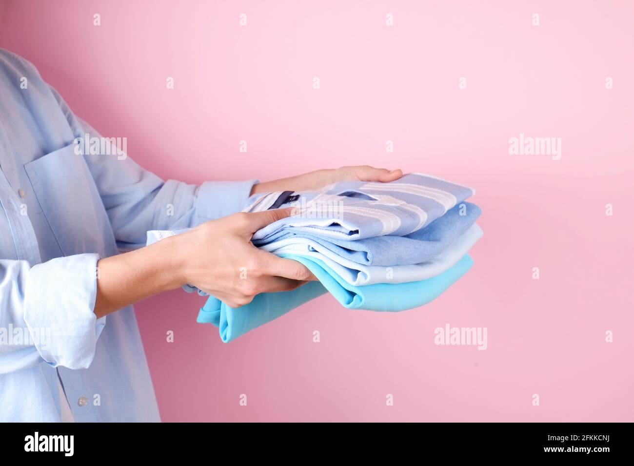 Primo piano di una giovane donna che indossa una camicia di cotone blu con una pila di camicie multicolore perfettamente piegate. Femmina con pile di indumenti di colore diverso in h Foto Stock