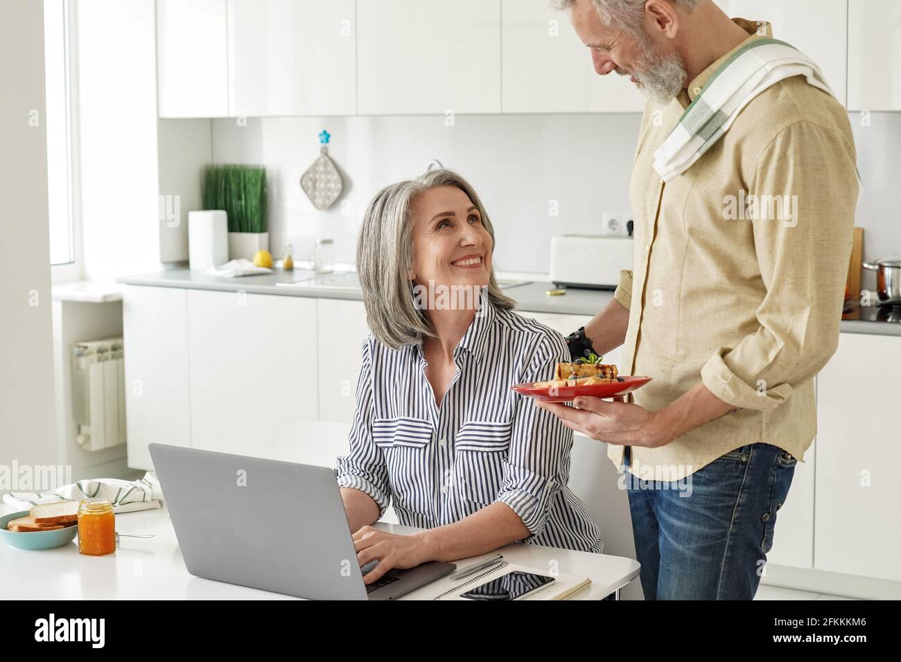 Marito anziano che prepara la colazione a una moglie felice che lavora dall'ufficio domestico. Foto Stock