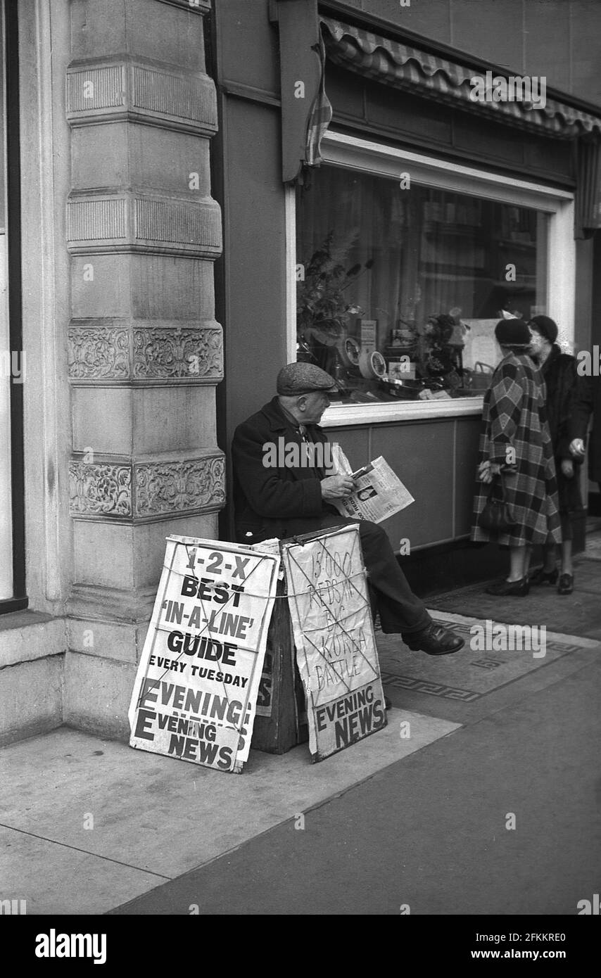 anni '50, hsitorical, un vecchio venditore di giornali maschili con carta in mano e indossando un cappello di stoffa seduto fuori su un pavimento. Il titolo di The Evening News, '15,000 Reds in Big Korea Battle', Londra, Inghilterra, Regno Unito. La guerra coreana si è svolta tra giugno 1950 e luglio 1953. Foto Stock