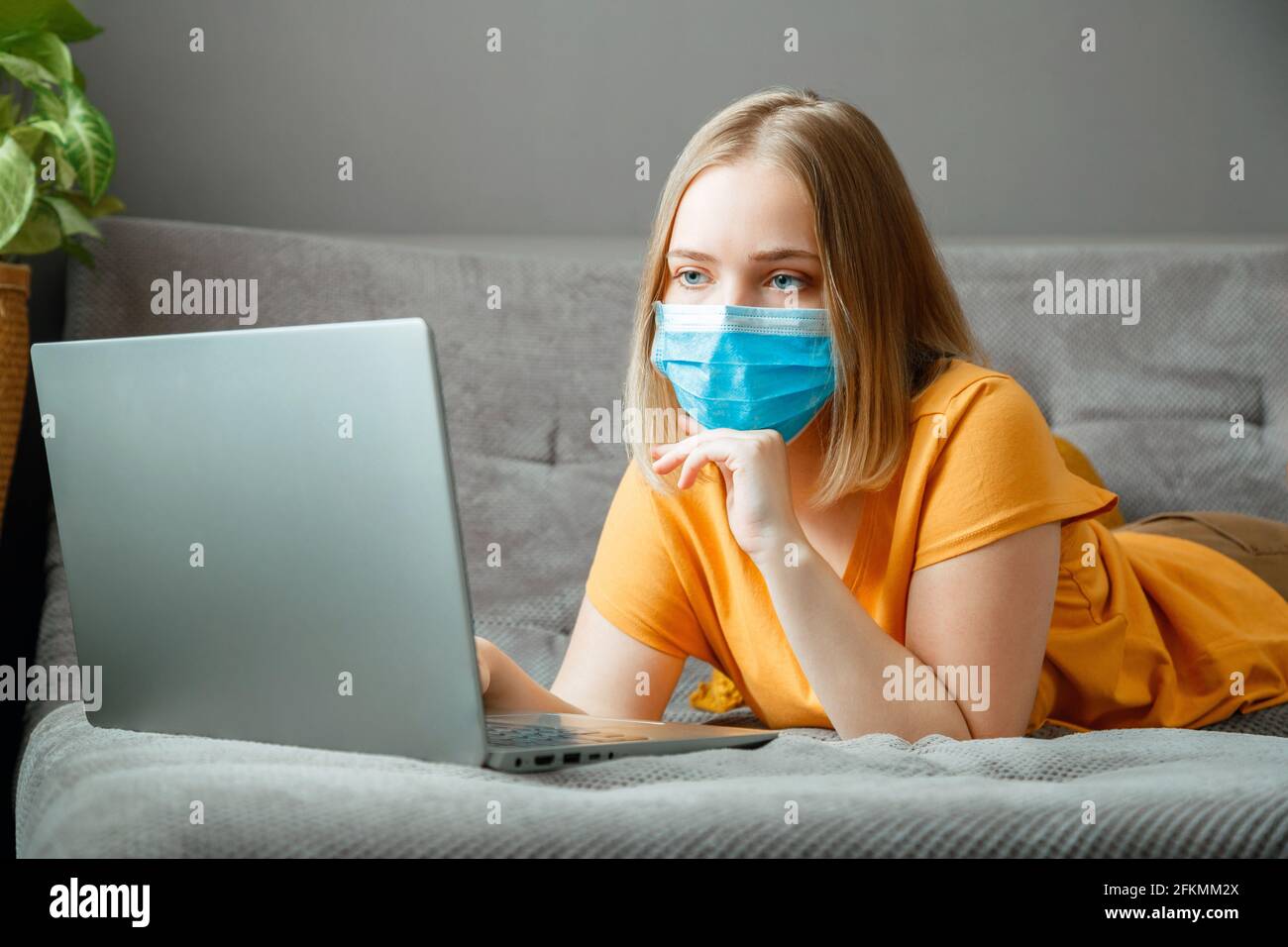 Donna in maschera medica di protezione lavoro utilizzando il computer portatile a casa ufficio si trova sul divano. Teen girl in mask formazione online via computer portatile covid 19 Foto Stock