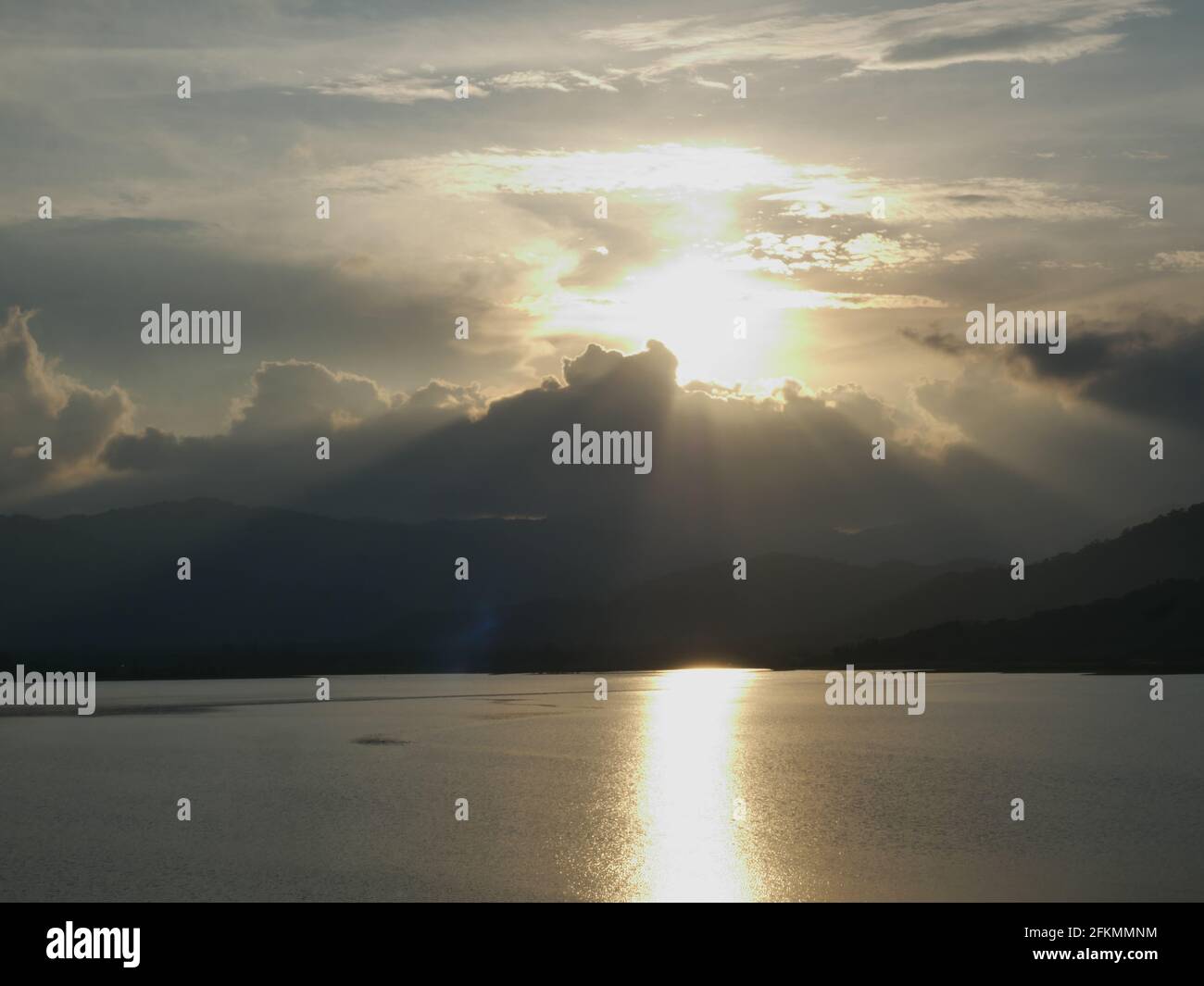 Il sole giallo dietro la sagoma della nuvola e della montagna si riflette sulla superficie del lago al tramonto, la superficie dell'acqua scintilla Foto Stock