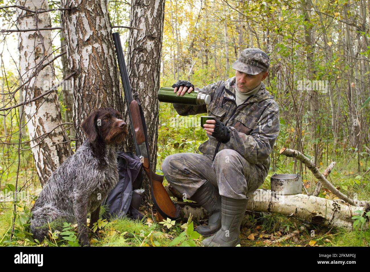 hunter seduto su un albero caduto e versa il tè da un thermos, seduto accanto al suo cane Foto Stock