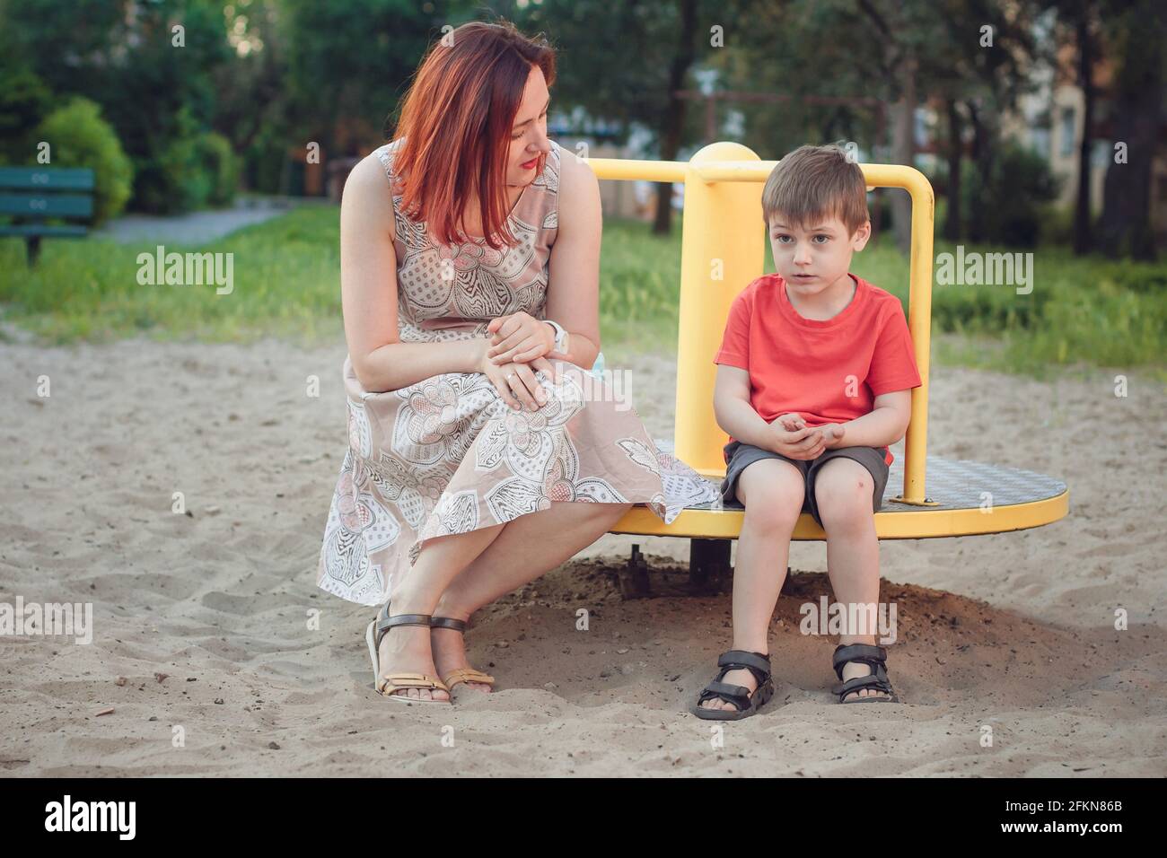 Mamma e figlio sul parco giochi. Ragazzo in T-shirt rosso brillante e giovane donna in abito all'aperto estate. Vita quotidiana. Concetto di infanzia e rapporto di Foto Stock