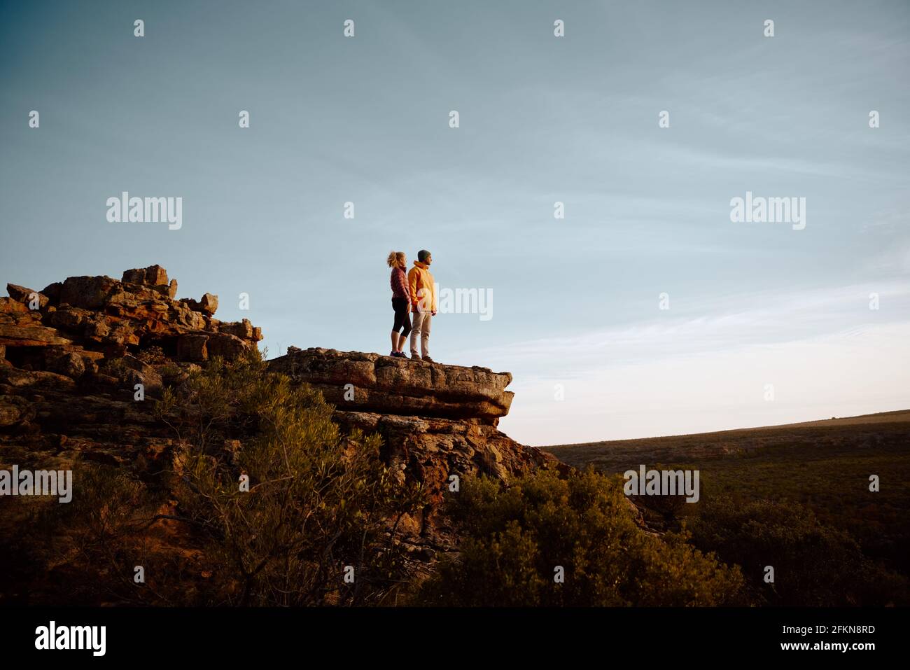 Vista ad angolo basso di un giovane uomo e di una donna che guarda in piedi con una splendida vista dopo aver fatto un'escursione in cima alla scogliera di montagna Foto Stock