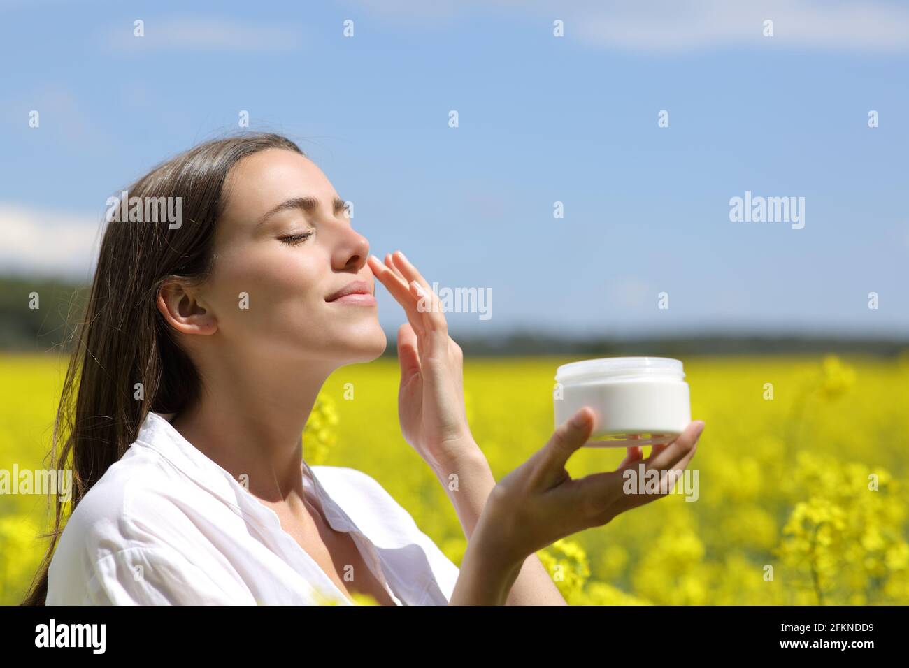 Donna di bellezza che tiene il vaso che applica la crema idratante sul viso dentro un campo fiorito Foto Stock