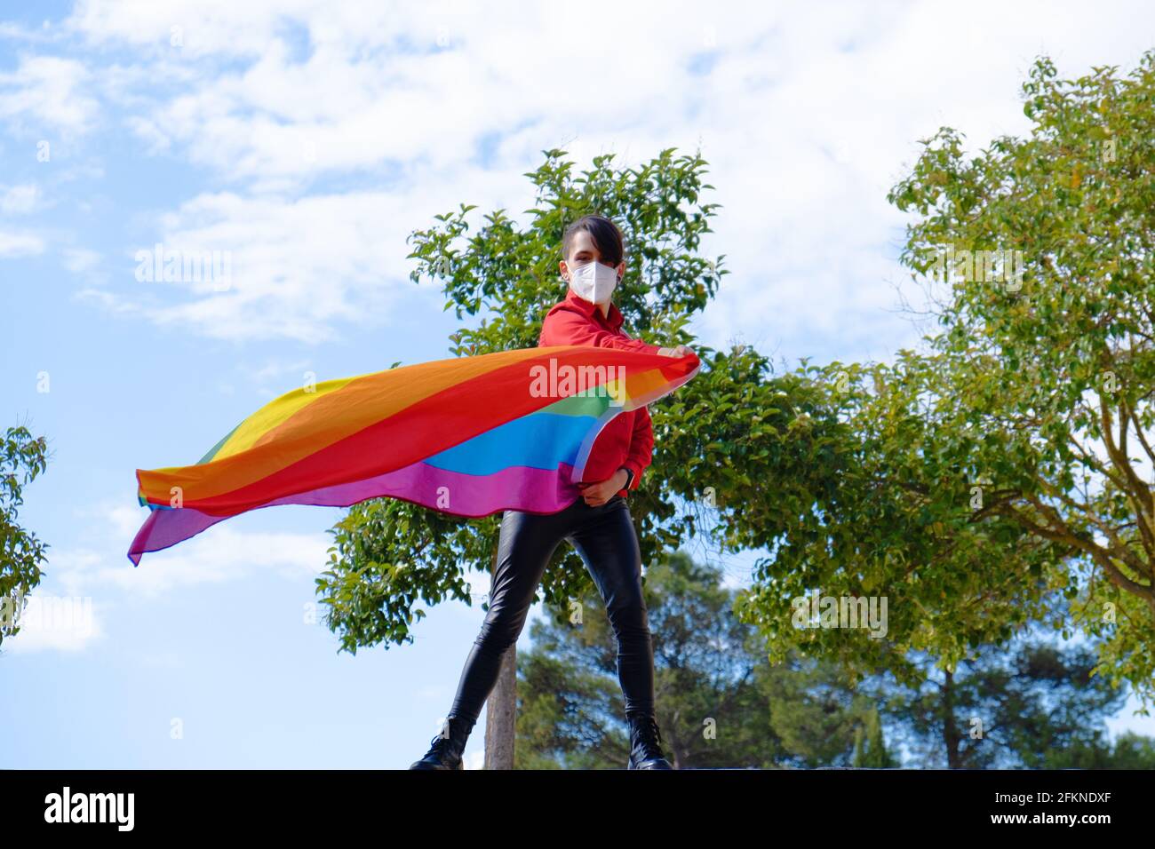 Una ragazza transgender con la bandiera gay e una maschera chirurgica. Persone autentiche non binarie. Gay Pride Day durante la pandemia di Covid Foto Stock