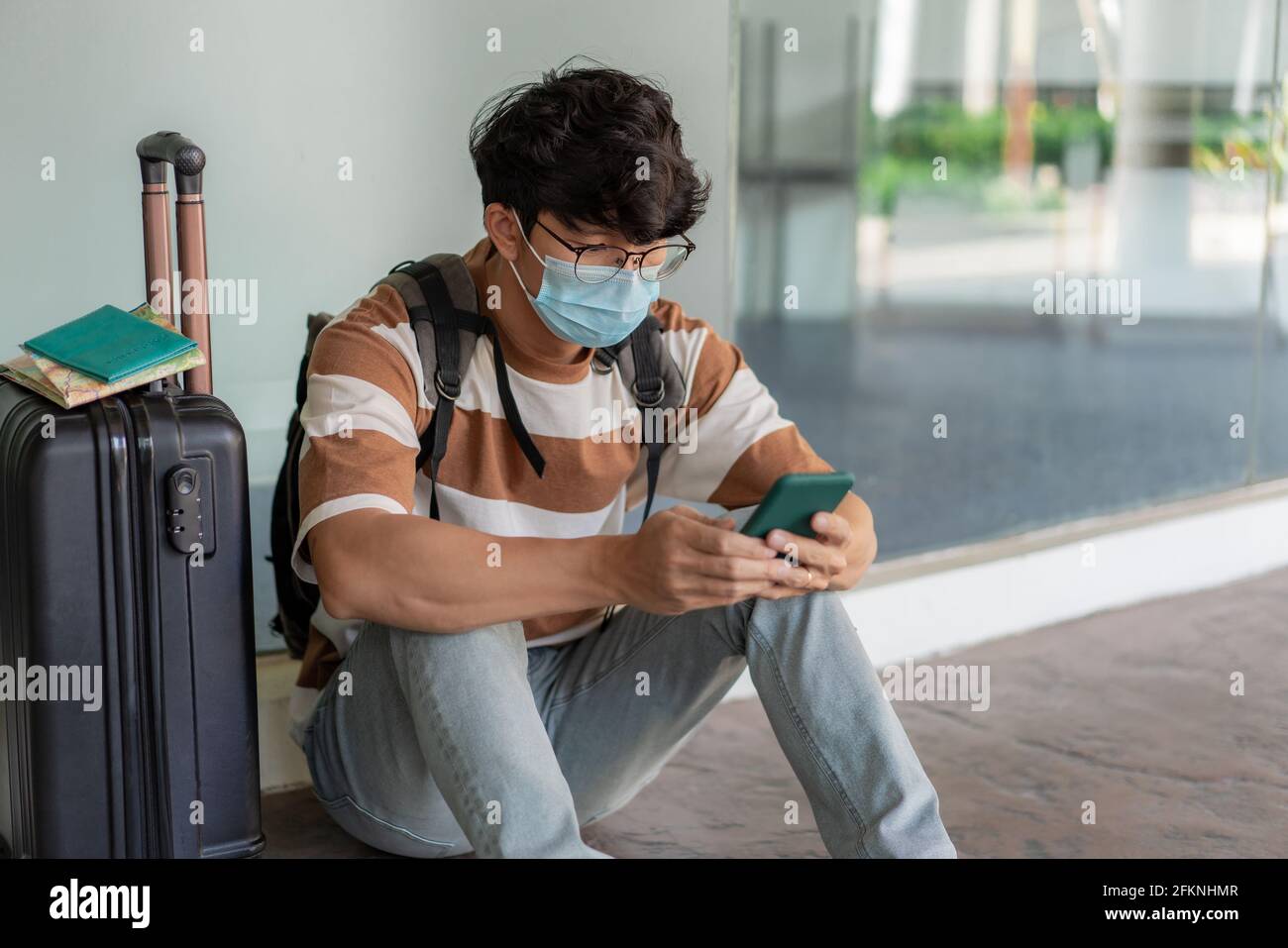 Viaggi. Un uomo che indossa occhiali con una camicia a righe e uno zaino, indossa una maschera, siediti sul pavimento. Utilizzando un telefono durante il viaggio con i bagagli da viaggio sul su Foto Stock
