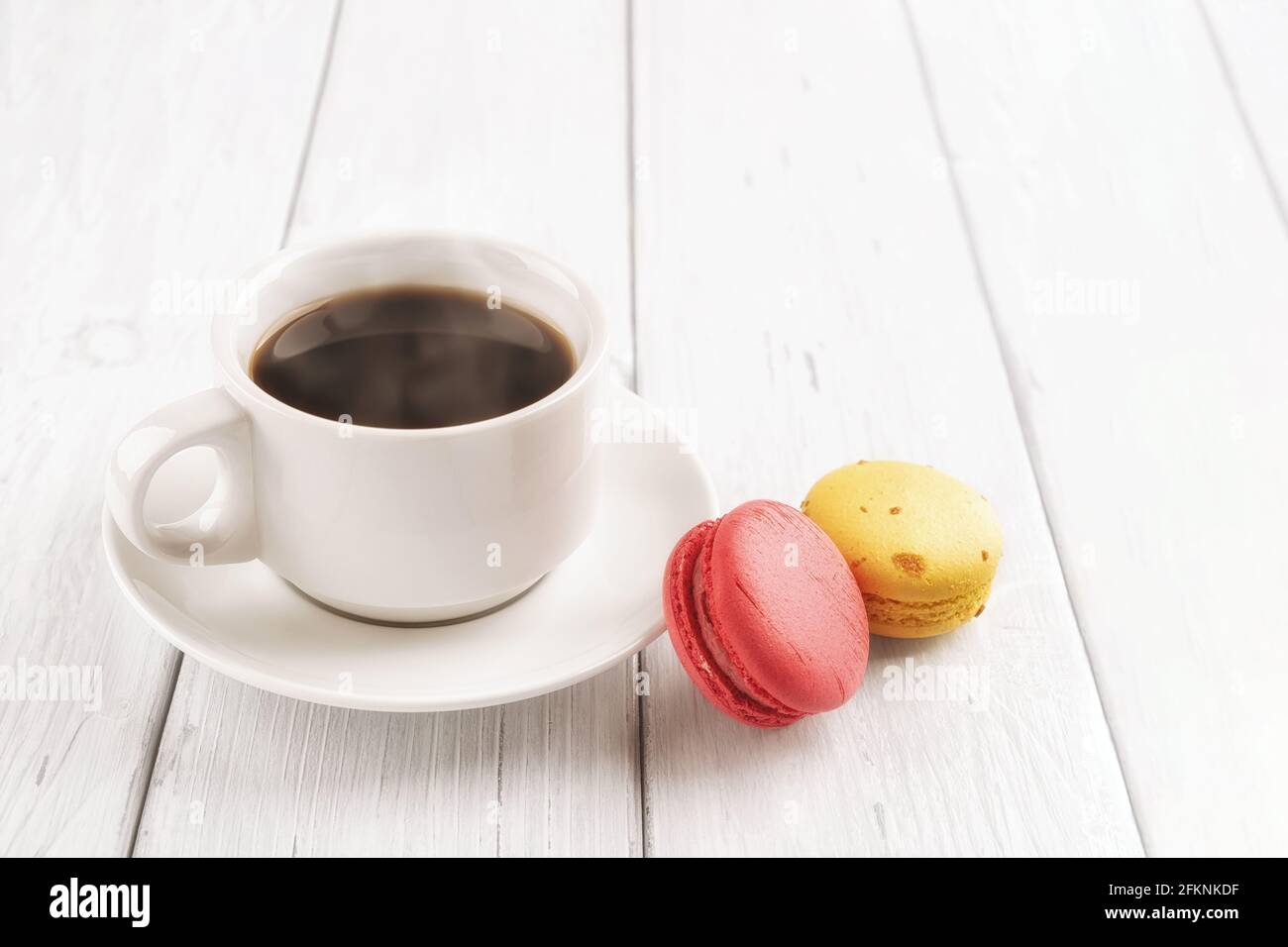 Tazza di caffè caldo e due macaroni su un tavolo di legno bianco. Colazione dolce con deliziosi biscotti alla meringa di mandorle. Immagine chiave alta. Foto Stock