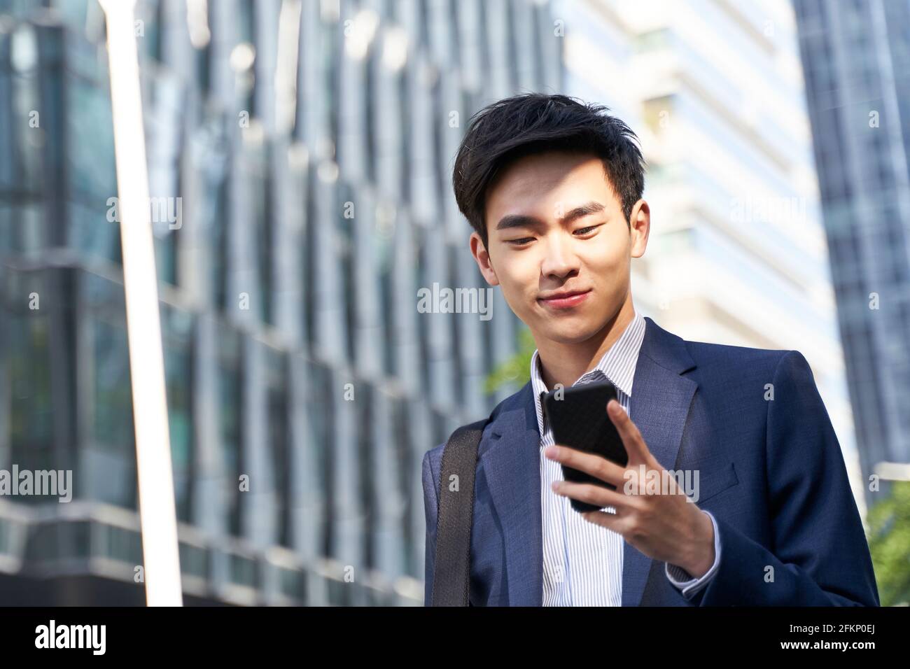 giovane uomo d'affari asiatico che guarda i messaggi sul cellulare mentre cammina nella strada nel centro della città moderna Foto Stock