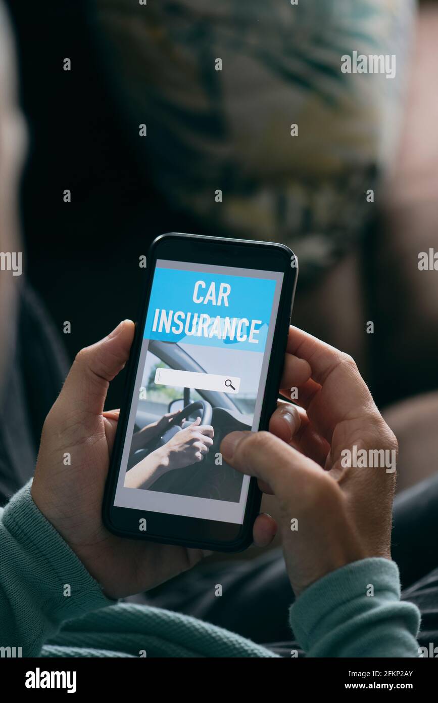 un giovane caucasico, seduto in soggiorno, cerca un'assicurazione auto online sul suo smartphone, con un motore di ricerca simulato Foto Stock