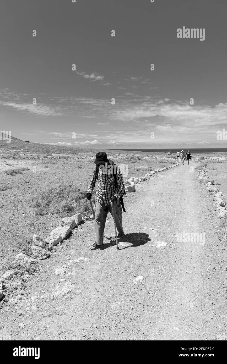 Uomo con maschera nera, cappello e camicia di controllo cammina con due bastoni lungo un percorso a la Tejita, Tenerife, Isole Canarie, Spagna Foto Stock