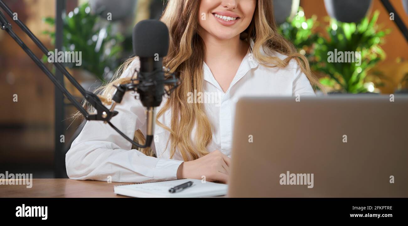 Primo piano di sorridente bella donna in camicia bianca che comunica online con microfono e laptop. Concetto di trasmissione live di processo per i seguaci AN Foto Stock