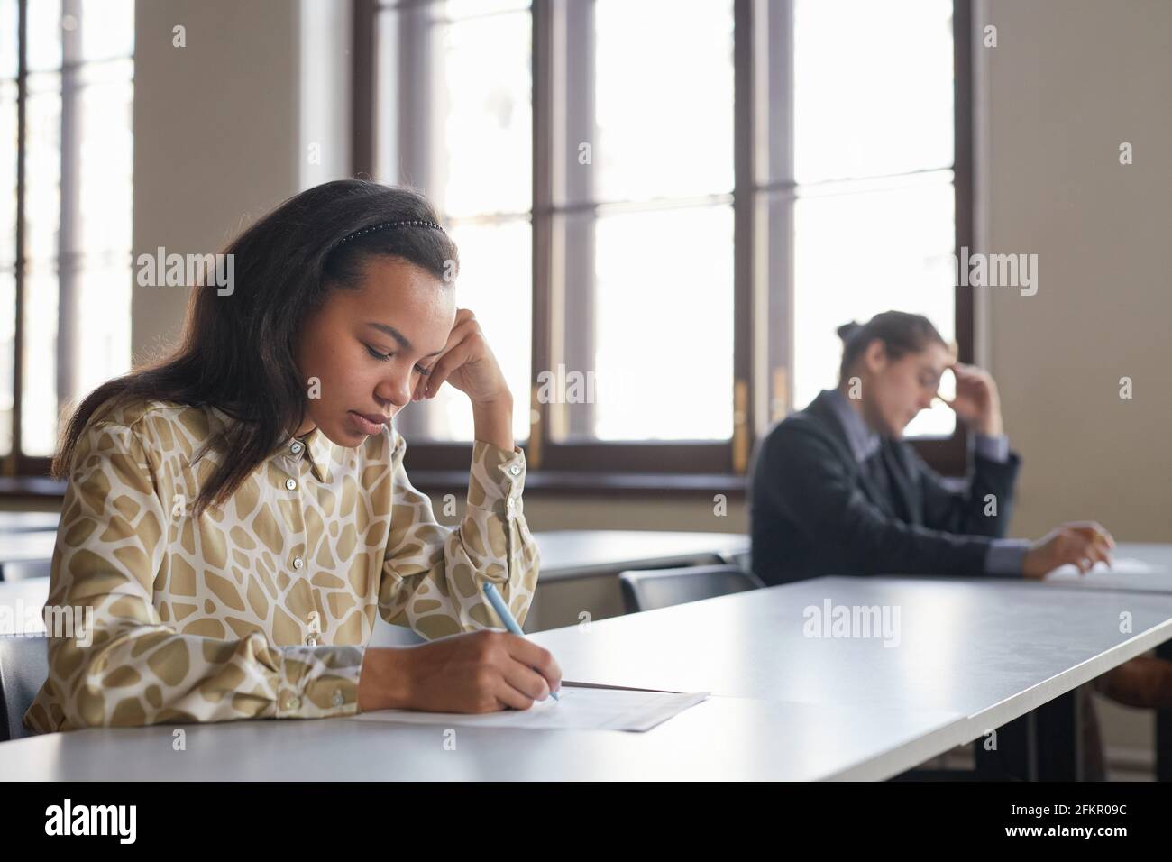 Vista laterale ritratto di due studenti che si stanno svolgendo esami di fila mentre si siedono alla scrivania con distanza sociale, concentrarsi sulla giovane donna afroamericana in foregroun Foto Stock