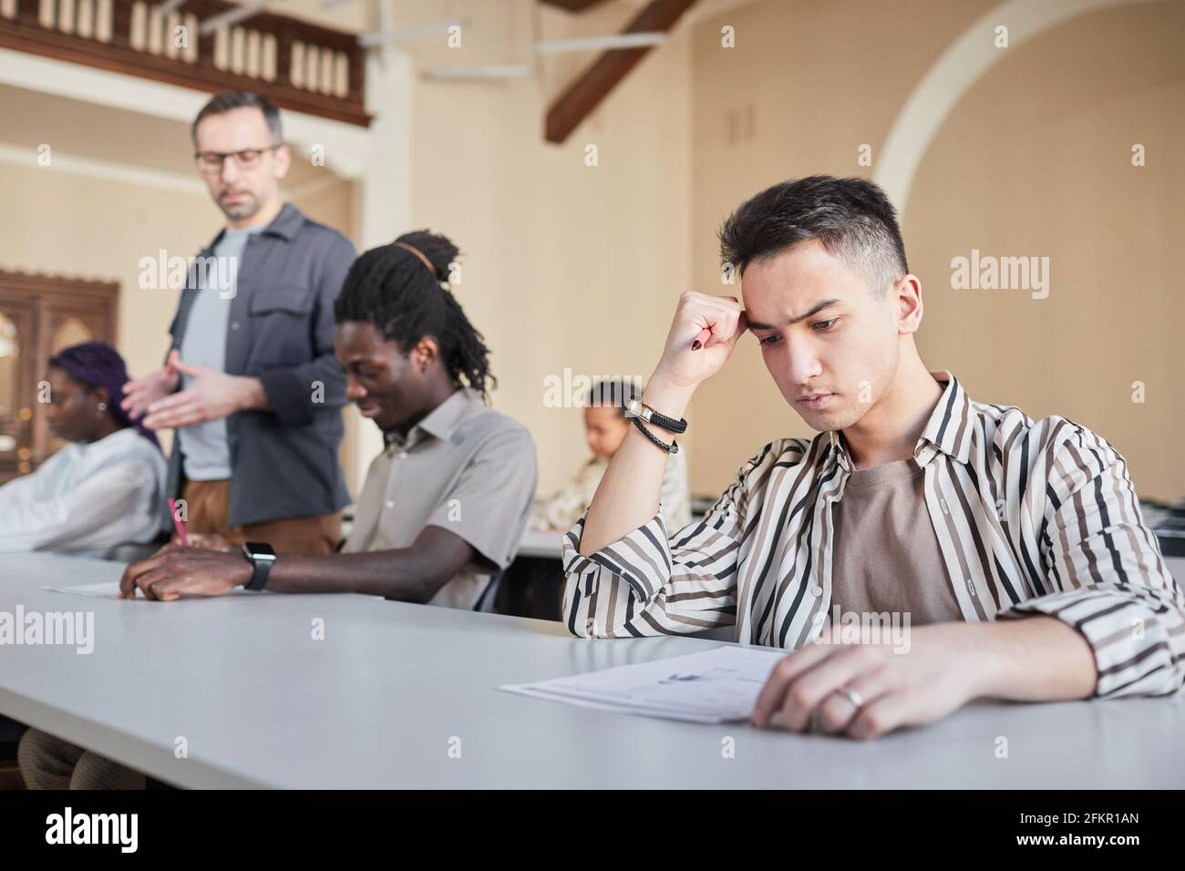 Ritratto degli studenti che si sottopone a un esame di fila mentre si siedono alla scrivania in auditorium, concentrarsi sul pensiero di un giovane asiatico in primo piano, copy space Foto Stock