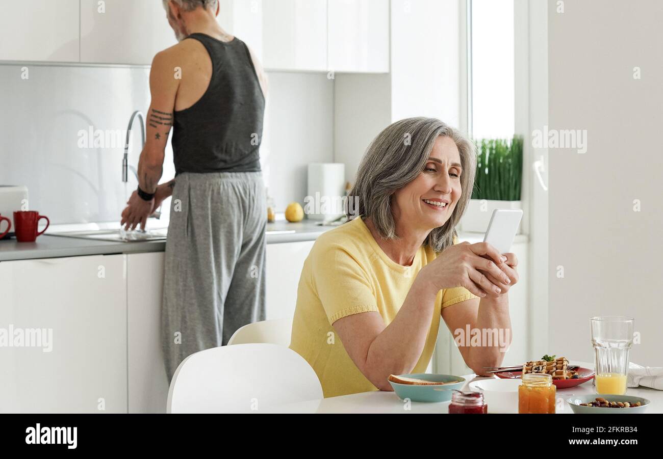 Felice moglie anziana che usa il telefono mentre il marito anziano lava i piatti in cucina. Foto Stock