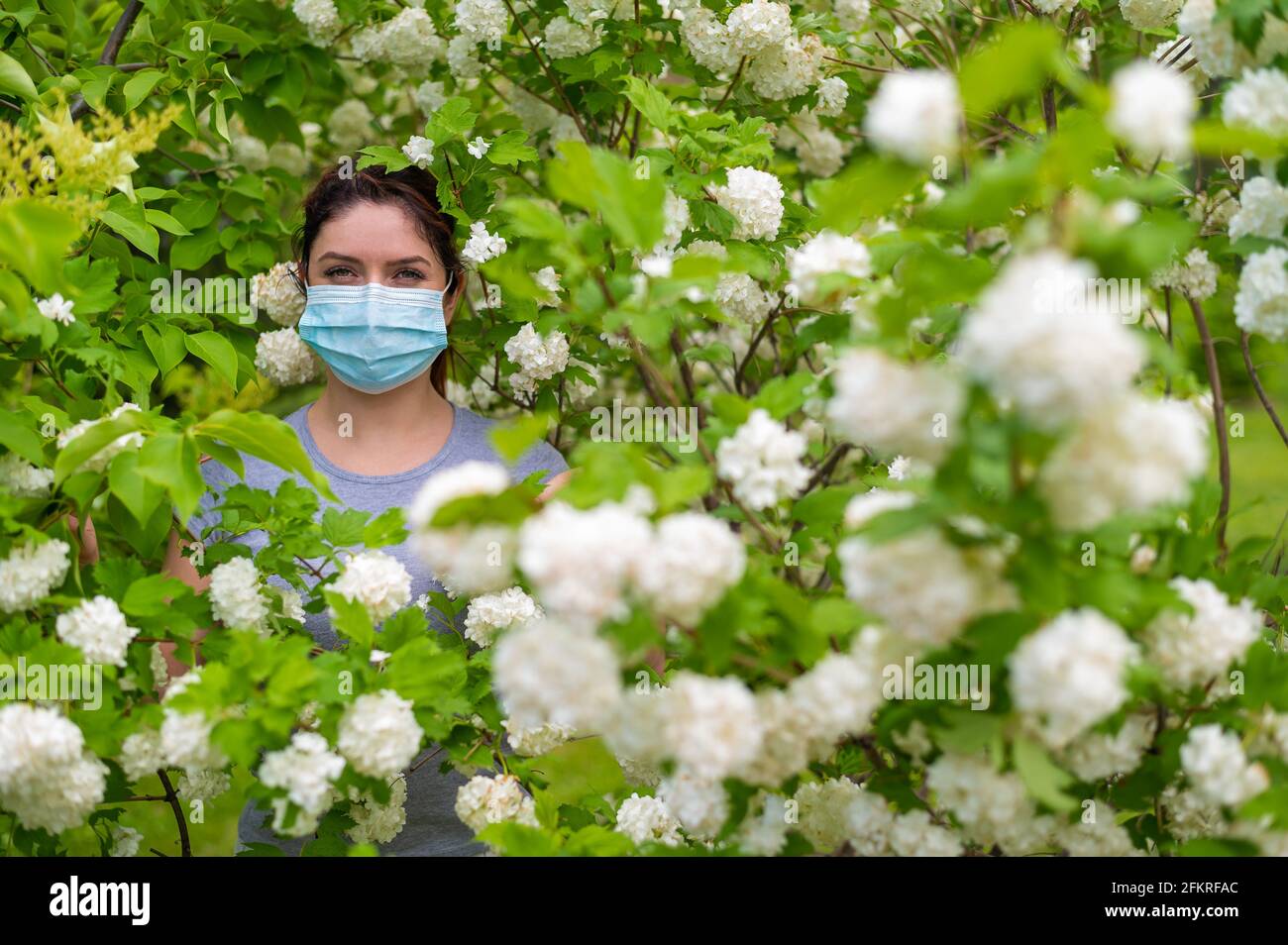 Una donna infelice dai capelli rossi in una maschera medica si trova accanto a un albero di mele in fiore in un parco. Rinite allergica stagionale. La ragazza soffre di un Foto Stock