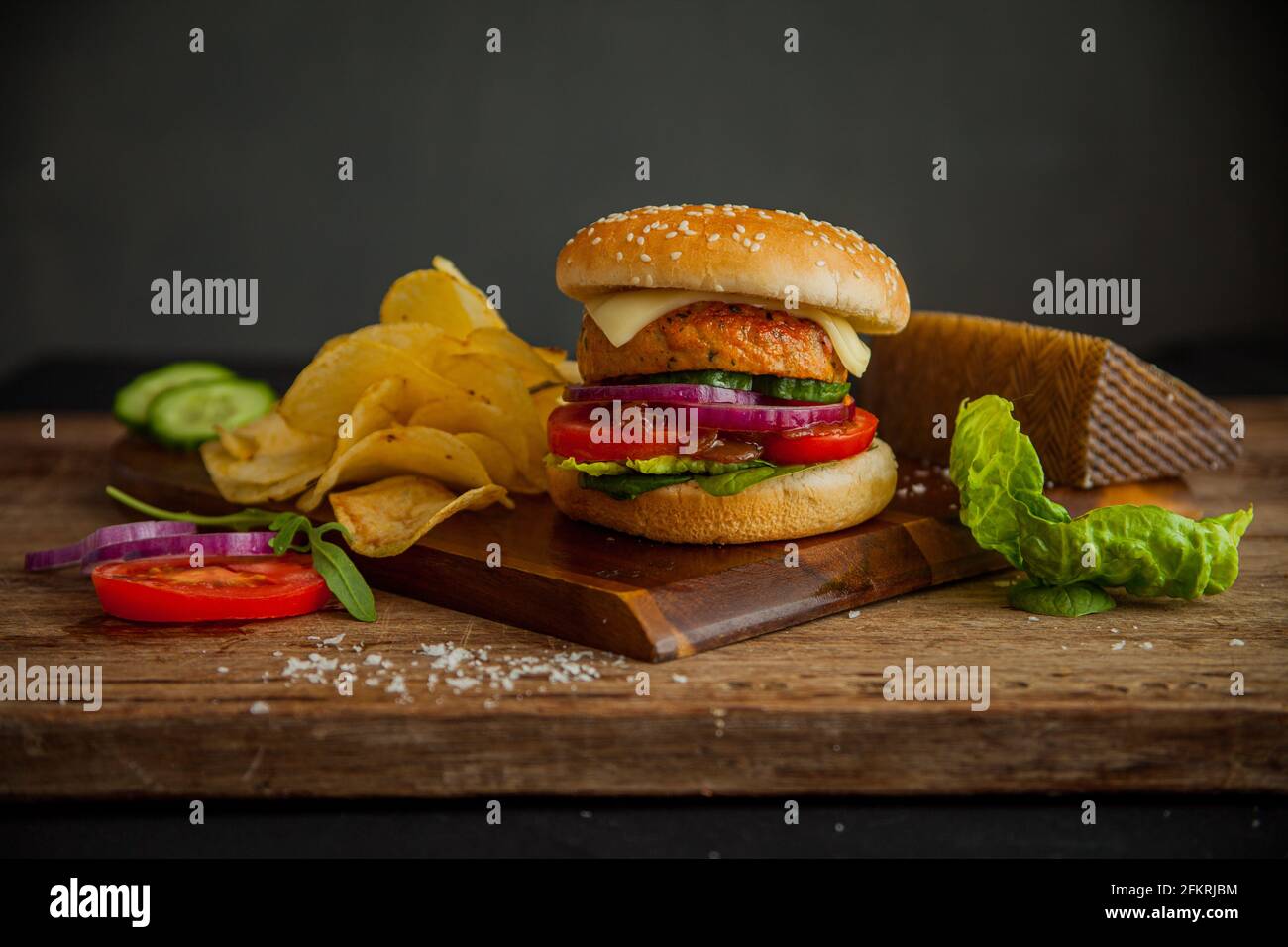 Hamburger di formaggio di pollo su tavola di legno con patatine, spicchi di formaggio e insalata con un semplice sfondo grigio scuro Foto Stock