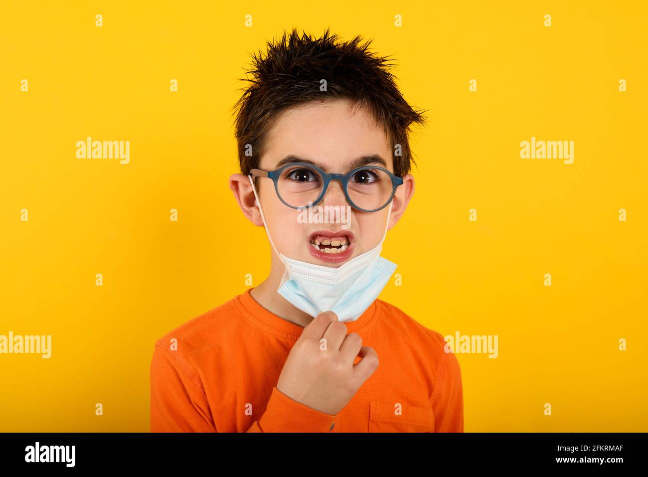 Ritratto di un bambino arrabbiato che vuole rimuovere maschera facciale per covid-19 coronavirus. Sfondo giallo Foto Stock