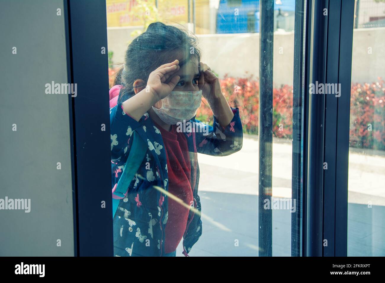 Studente elementare o asili che indossa una maschera protettiva che guarda verso l'interno attraverso la finestra della porta della scuola Foto Stock