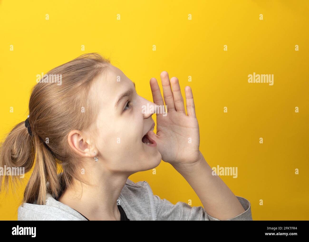 Primo piano di ritratto di giovane ragazza agitata con capelli tirati indietro in coda di pony, urlando forte, con mano vicino al viso su sfondo giallo. Chiamata, lo Foto Stock