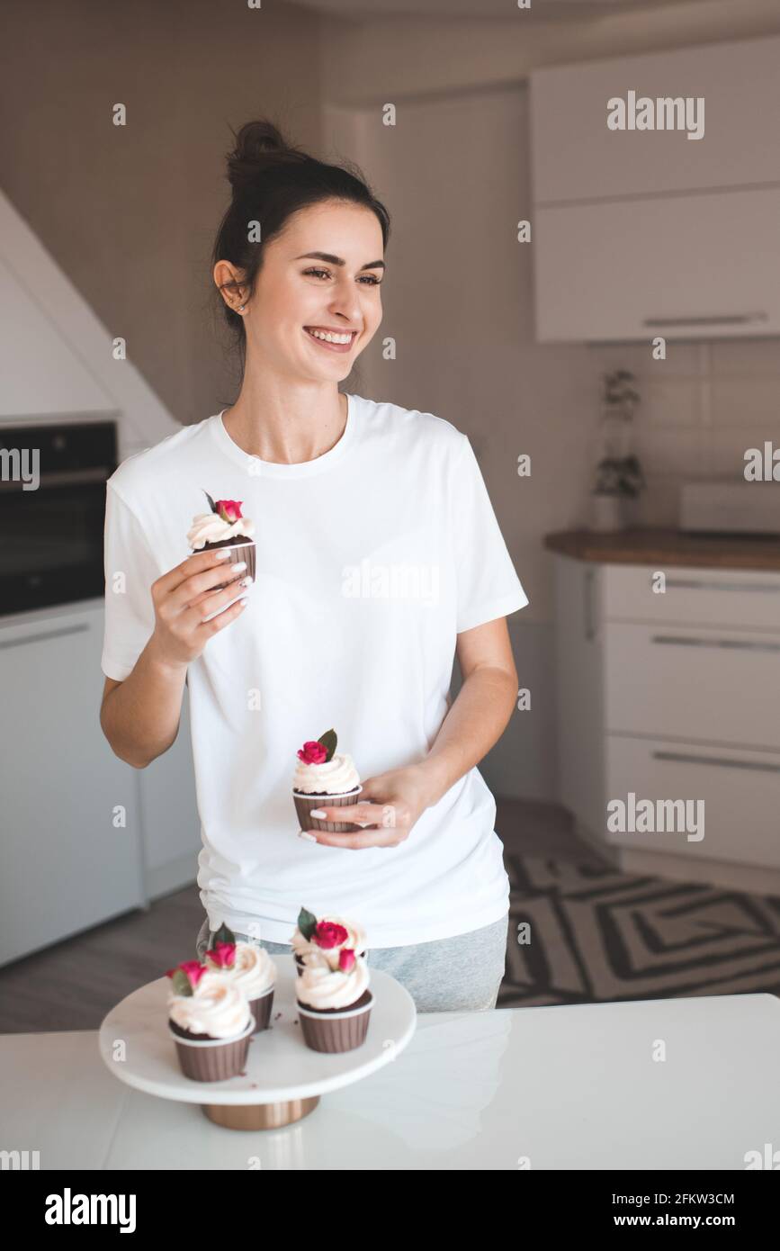 Donna felice che indossa abiti casual che reggono cupcakes al forno con formaggio crema in primo piano. Sorridente giovane ragazza adulta di 24-26 anni mangia gustosa pasticceria Foto Stock