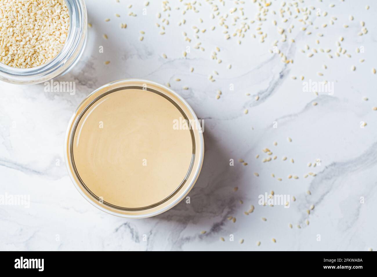 Pasta di sesamo - tahini in un vaso di vetro, sfondo di marmo bianco. Concetto di cucina israeliana. Foto Stock