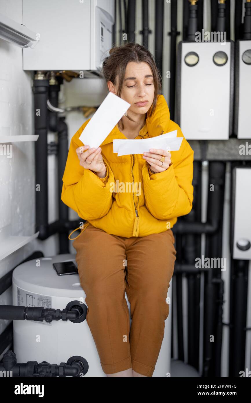 Donna confusa guarda le bollette elettriche, confrontando i dati con il misuratore nel quadro elettrico a casa. Concetto di spese per la famiglia Foto Stock