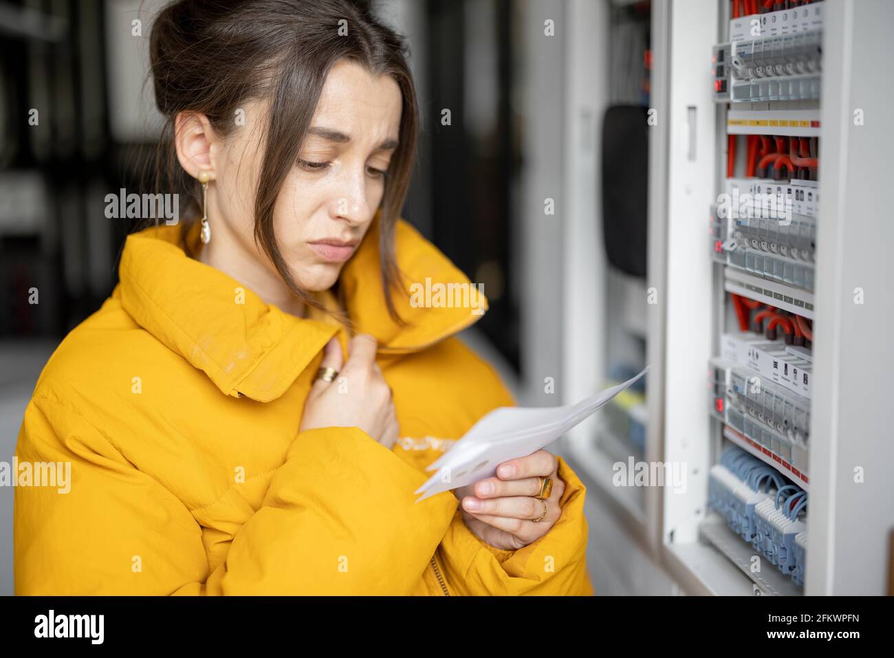 Donna confusa guarda le bollette elettriche, confrontando i dati con il misuratore nel quadro elettrico a casa. Concetto di spese per la famiglia Foto Stock