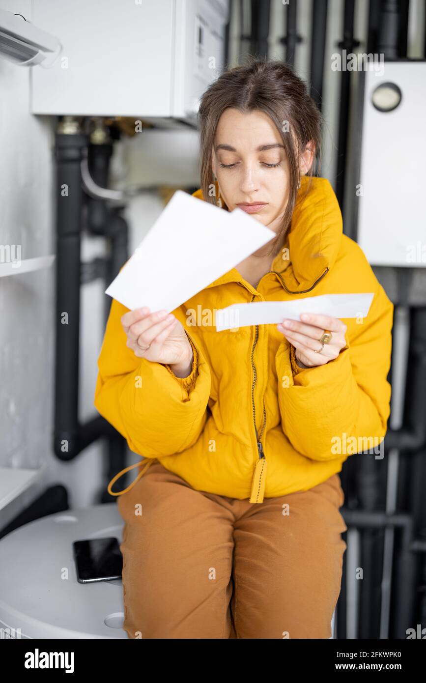 Donna confusa guarda le bollette elettriche, confrontando i dati con il misuratore nel quadro elettrico a casa. Concetto di spese per la famiglia Foto Stock