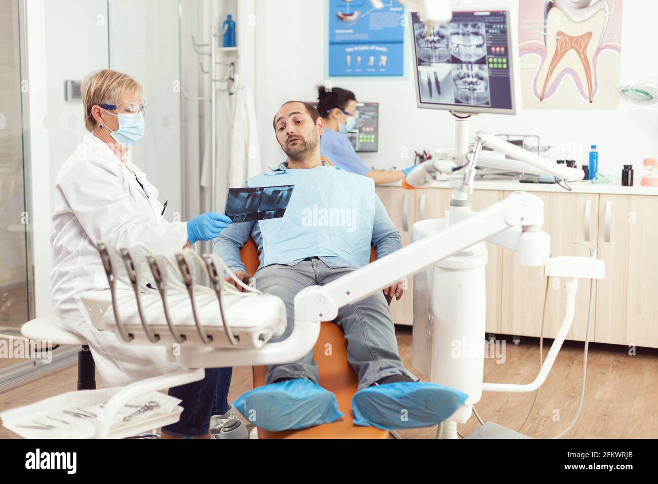 Donna anziana dentista trattamento malato paziente uomo in moderno studio di odontoiatria stomatologica mostrando la radiografia dei denti spiegare l'intervento dentale. In background infermiera che prepara il trattamento del mal di denti Foto Stock