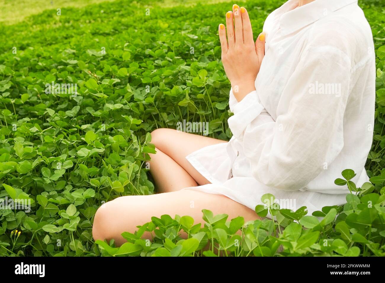 Irriconoscibile giovane donna che pratica yoga, seduta in posa di namaste Padmasana Lotus. Donna sportiva, camicia in cotone oversize che medita all'aperto, erba f Foto Stock