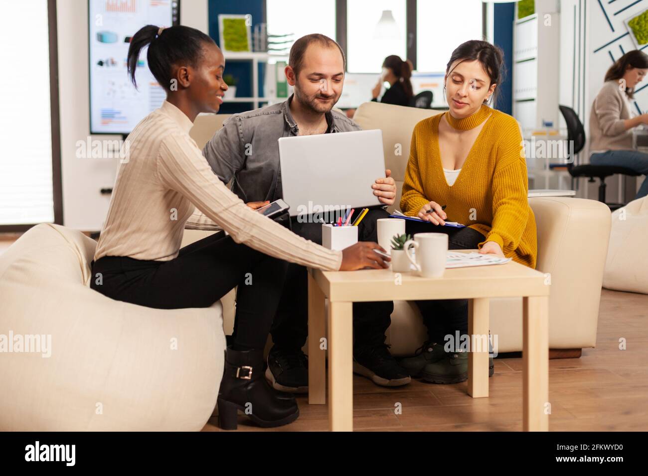 Riunione del team Startup con il leader aziendale che indica la strategia sui report finanziari. Lavoro di squadra in un ufficio moderno di diversi gruppi di colleghi internazionali seduti sul divano al tavolo nella gestione delle startup Foto Stock