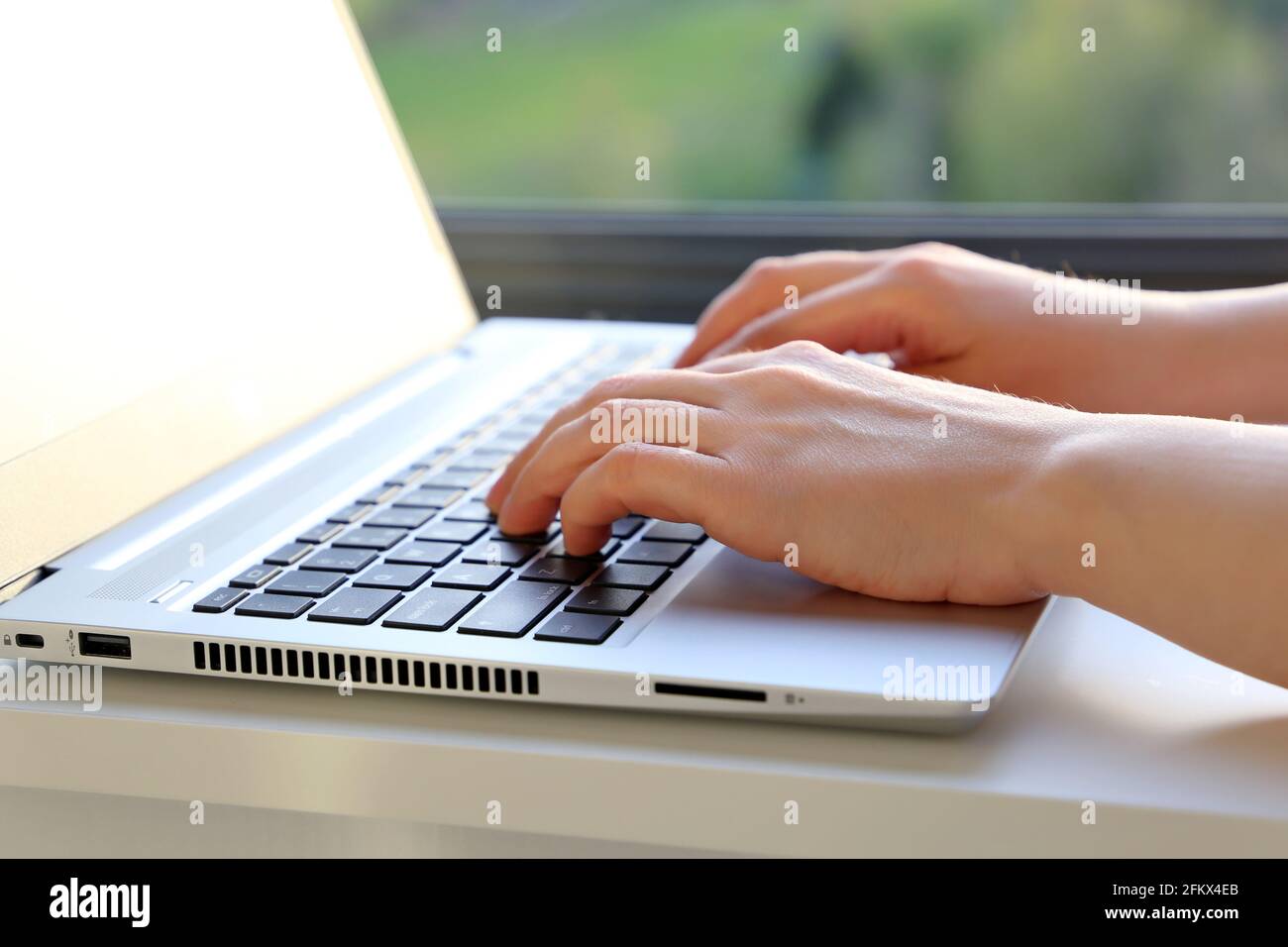 Le mani femminili sul laptop, le donne che si trovano sulla tastiera seduta vicino alla finestra con vista della natura. Ambiente di lavoro accogliente, concetto di lavoro in ufficio o a casa Foto Stock