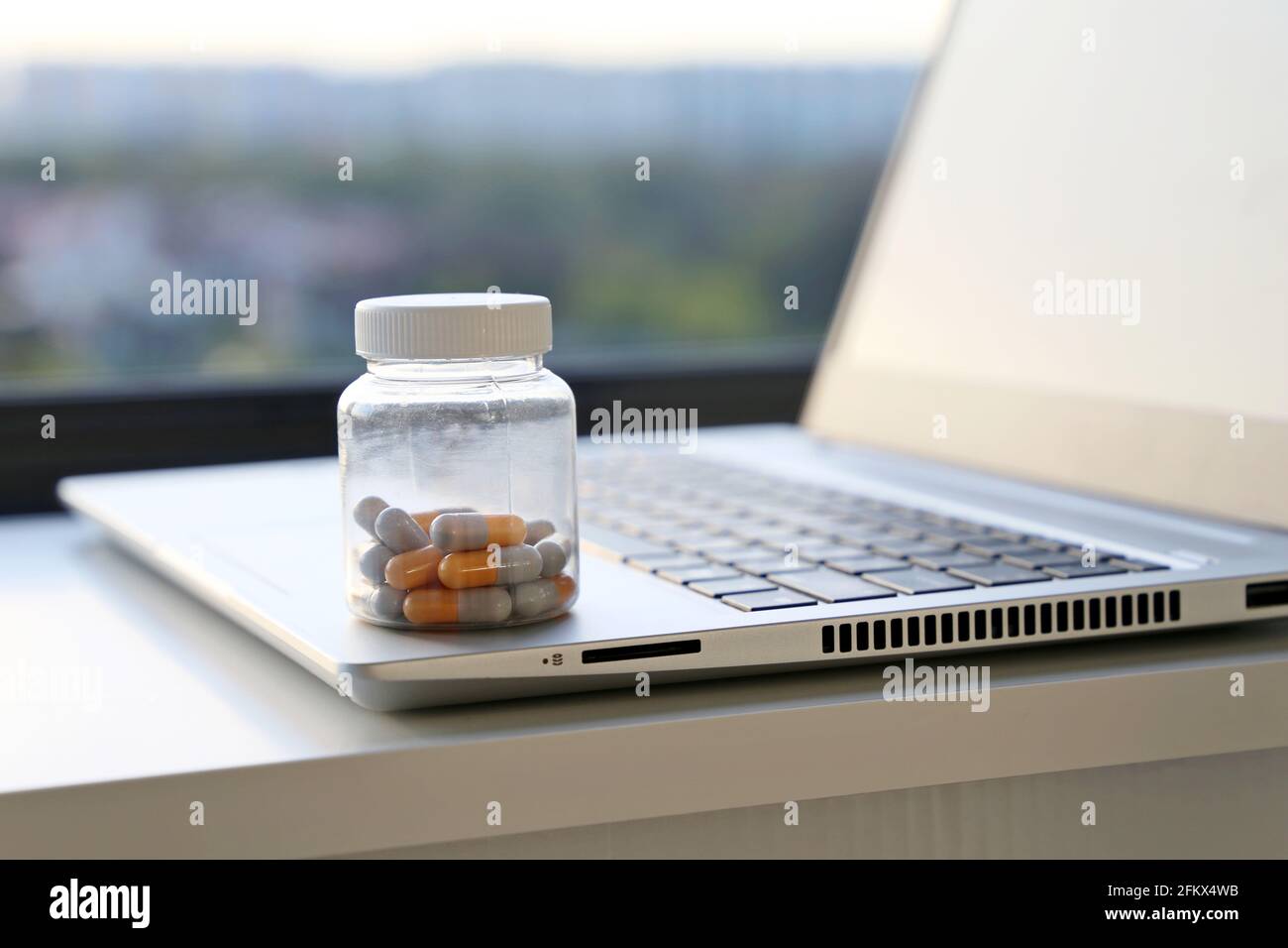 Pillole sulla tastiera del laptop sullo sfondo della finestra, capsule in un flacone. Concetto di assunzione di farmaci in ufficio, vitamine per l'energia e il lavoro Foto Stock