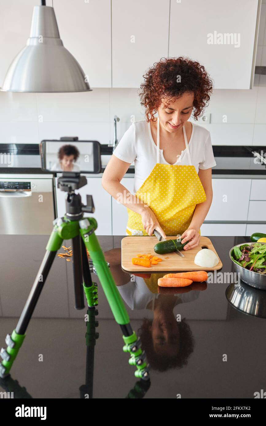 La donna youtuber si registra con il suo smartphone in cucina mentre si prepara una ricetta di insalata, c'è luce naturale e lei indossa una a gialla Foto Stock