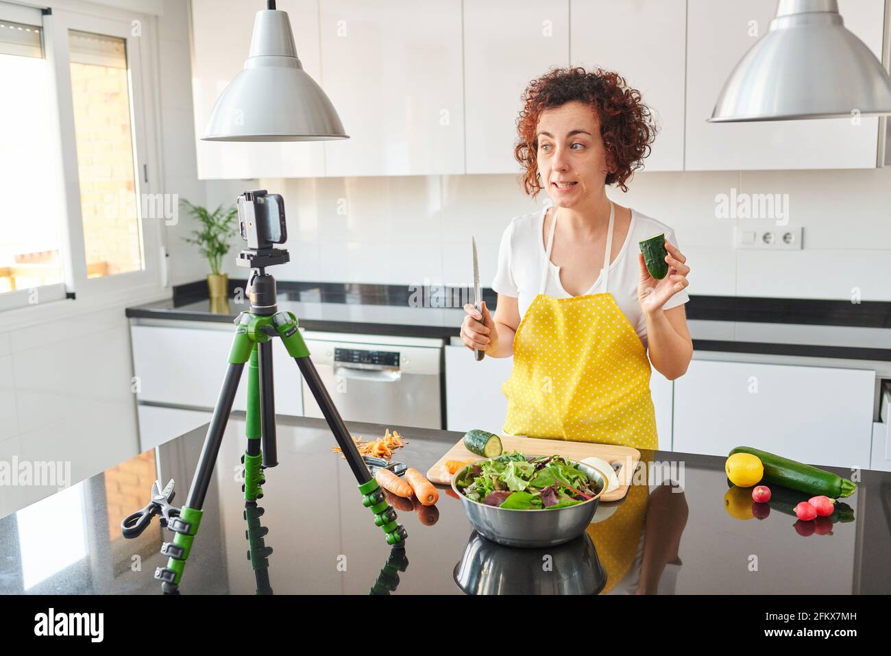 La donna youtuber si registra con il suo smartphone in cucina mentre si prepara una ricetta di insalata, c'è luce naturale e lei indossa una a gialla Foto Stock
